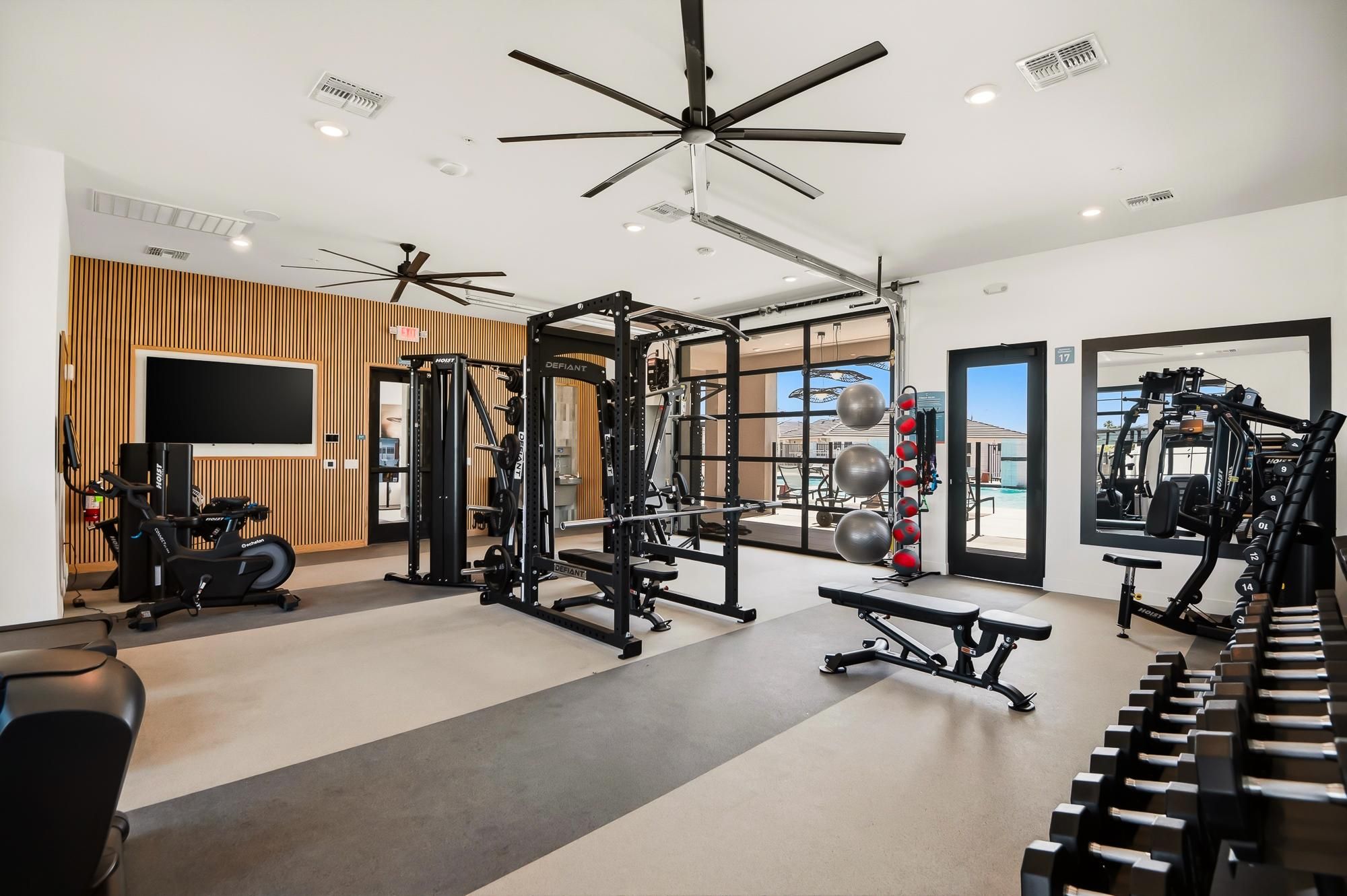 Modern gym with various exercise machines, free weights, medicine balls, and large windows letting in natural light.
