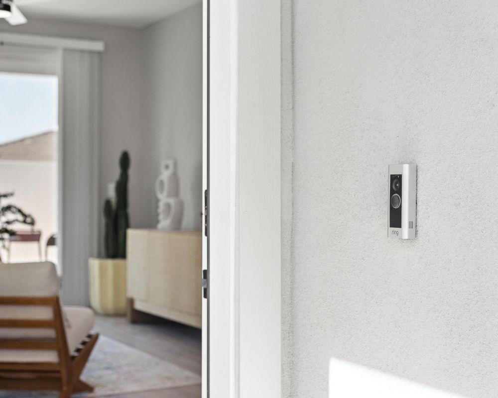 Verge Apache Junction Ring doorbell is mounted on a white wall near the entrance of a modern, sunlit living room.