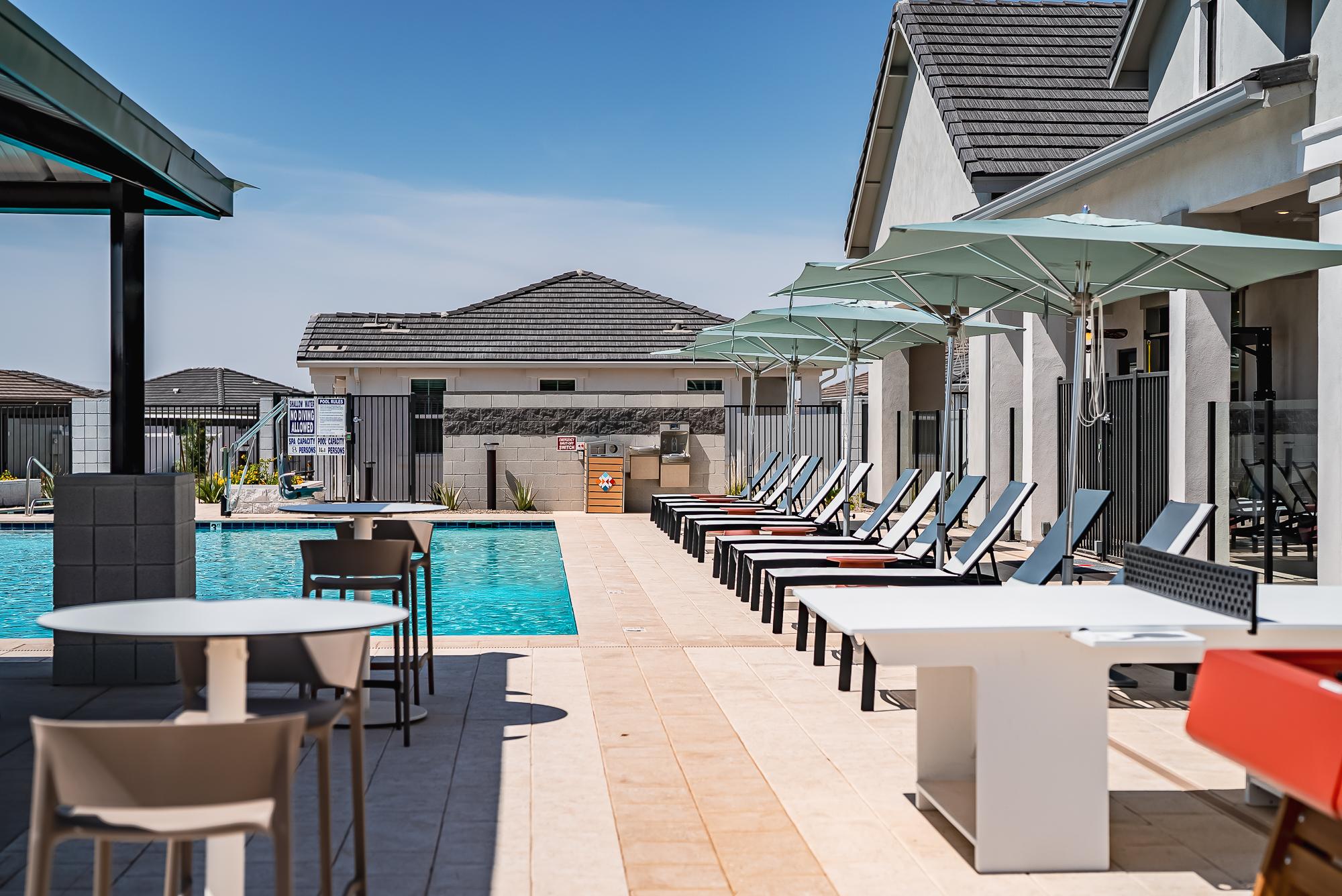 Outdoor pool area with lounge chairs, tables, and umbrellas next to modern buildings under a clear sky.