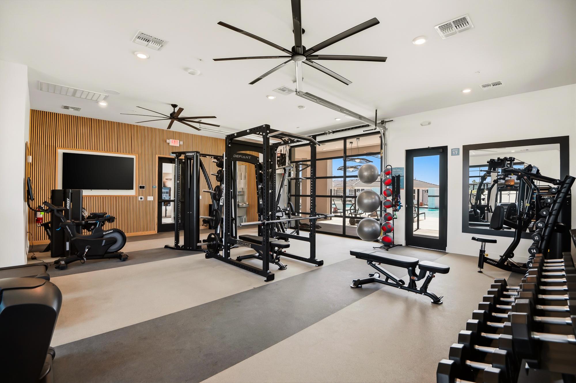 Modern gym with various exercise machines, free weights, medicine balls, and large windows letting in natural light.
