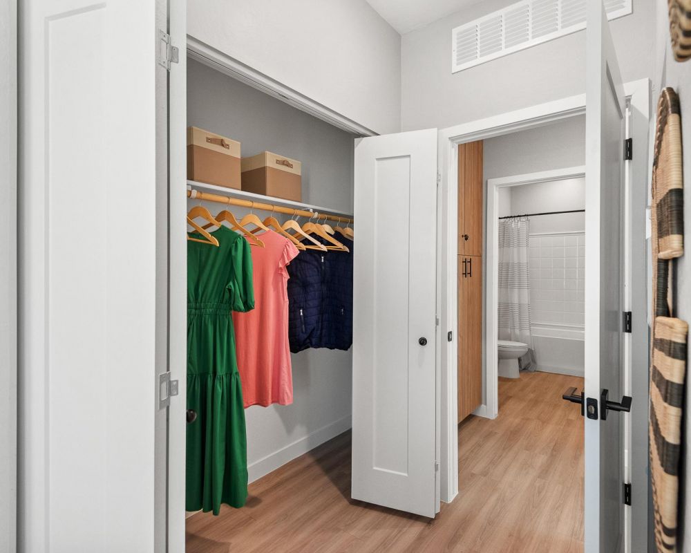 Verge Apache Junction walk-in closet with clothes on wooden hangers, boxes on a shelf, and a view into a bathroom.