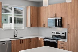 Modern kitchen with wooden cabinets, white countertops, a stove, microwave, sink, and window with blinds.